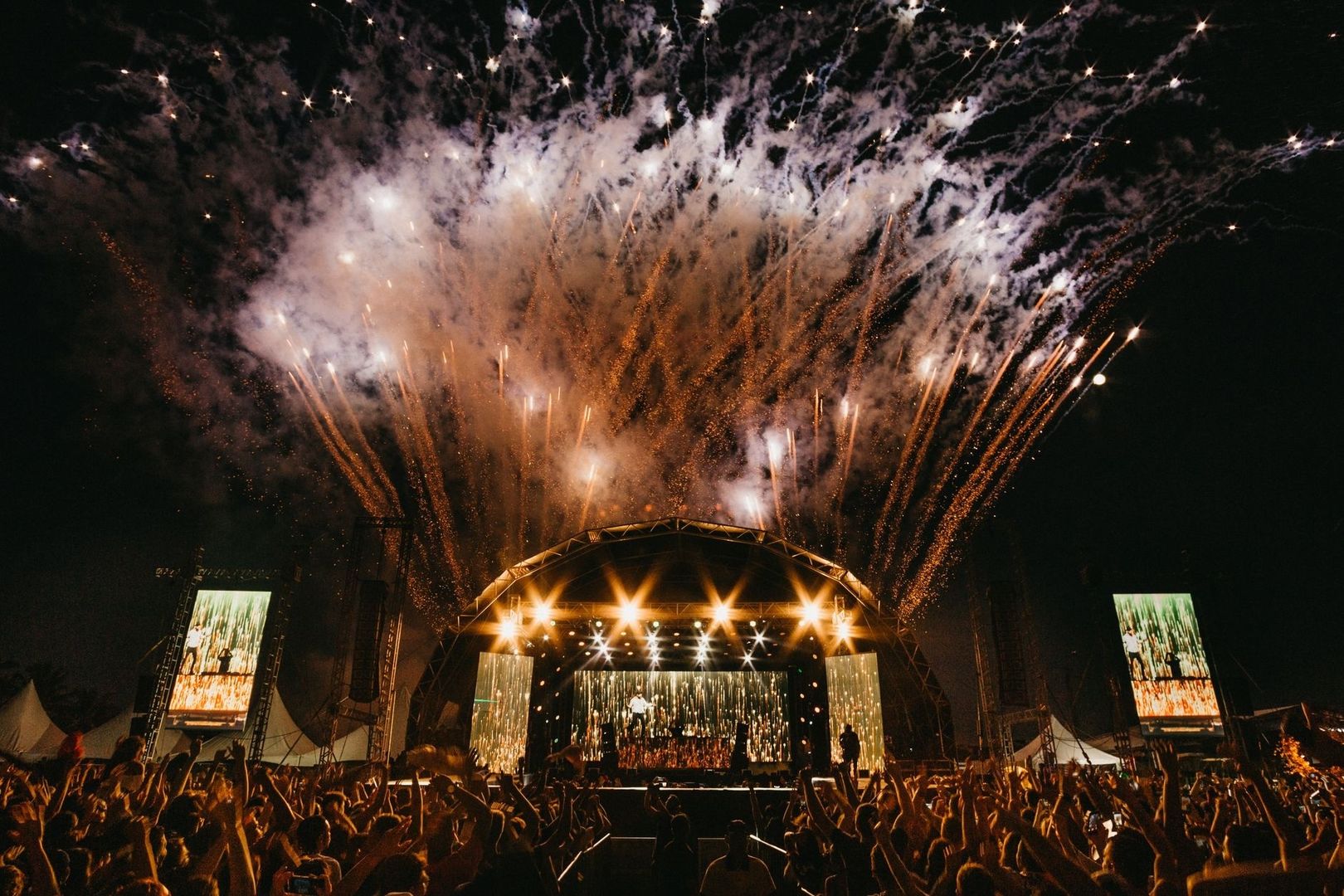 Crowded music festival stage illuminated by an expansive fireworks display